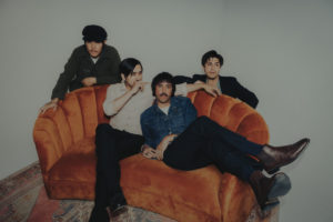 Four young men lounging on or squatting behind an orange couch