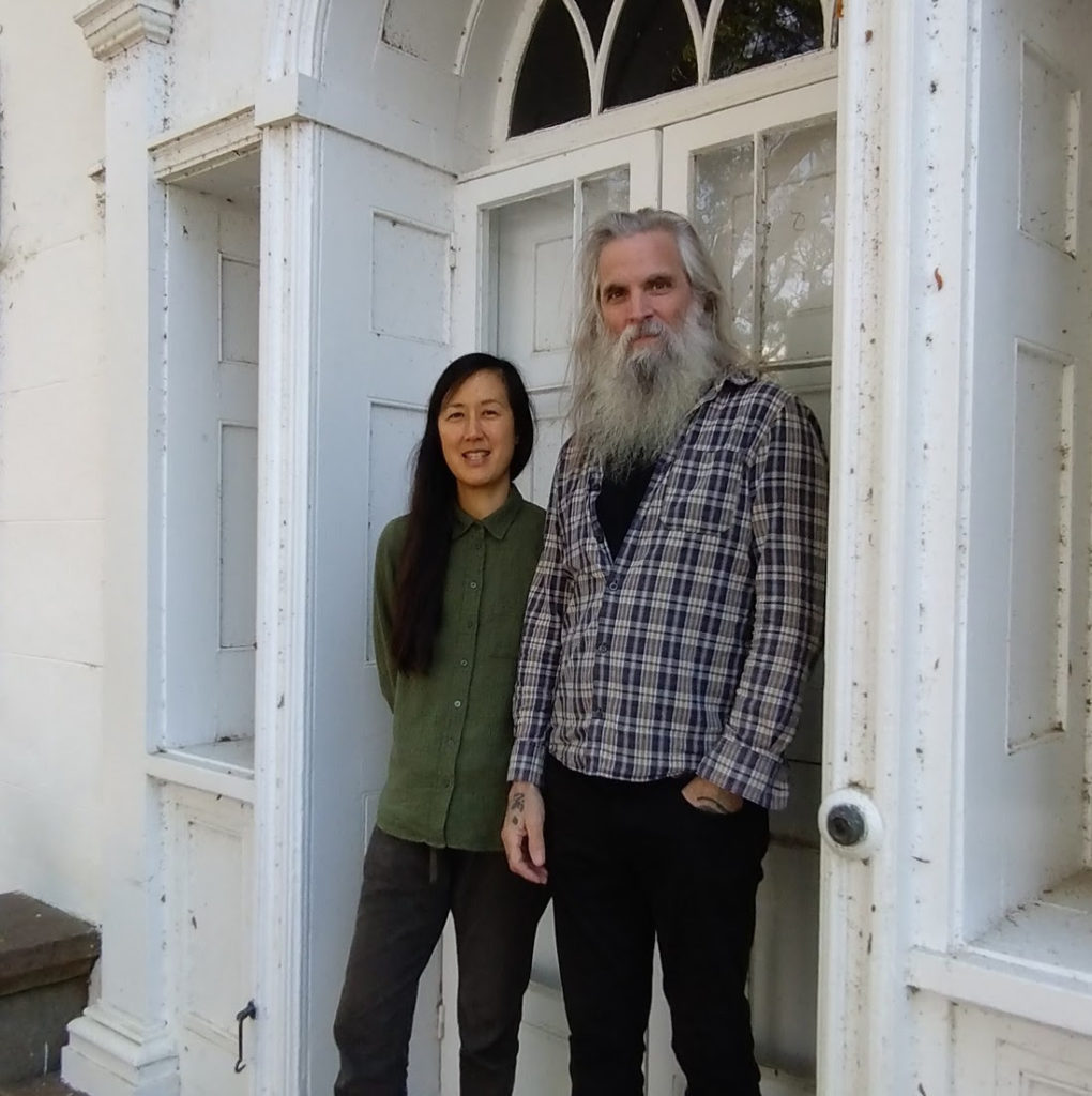 Tall man in flannel shirt with long grey hair and grey beard with Asian woman standing next to him in front of the door of a white house.