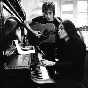 Black and white photo of Yoko Ono playing the piano with John Lennon standing beside playing the acoustic guitar