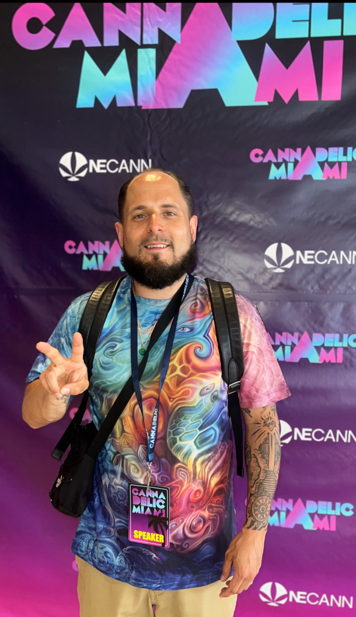 Bald man with beard wearing tie-dyed shirt giving peace sign in front of Cannadelic Miami background