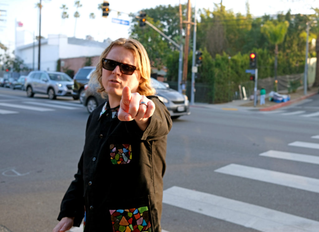 Ty Segall wearing dark clothes and pointing at the camera while crossing a busy street