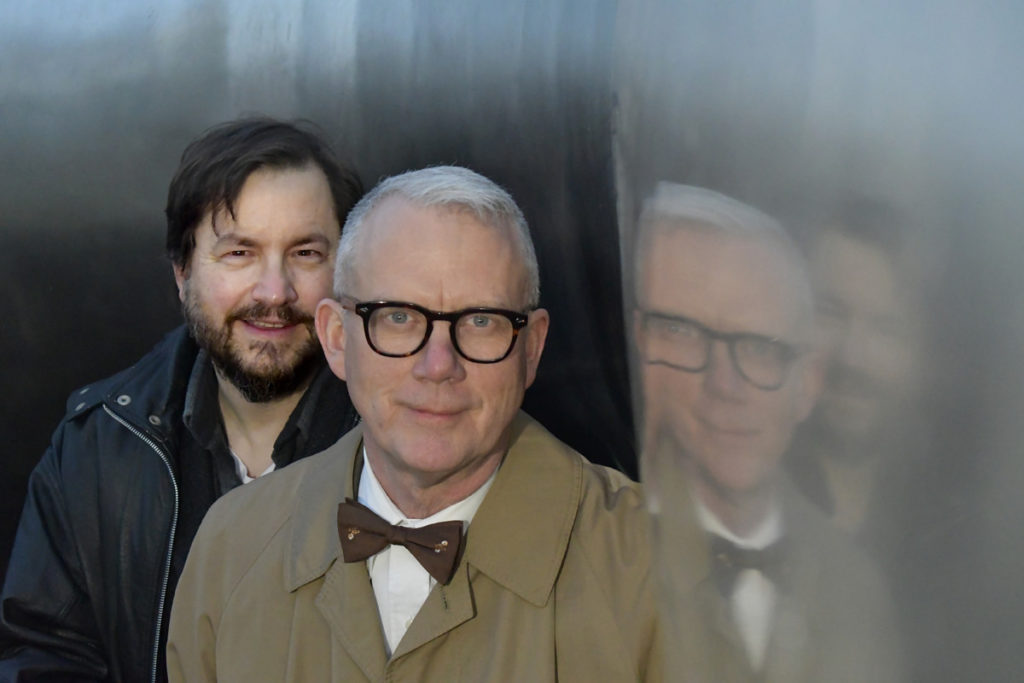 Close up of two men--the one with white hair and a bowtie in front with the dark-haired man standing right behind him next to the metal wall of the cylinder