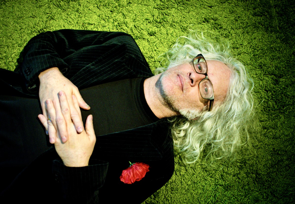 Man with long, white hair and a black, long-sleeved shirt lying in the green grass with his hands together