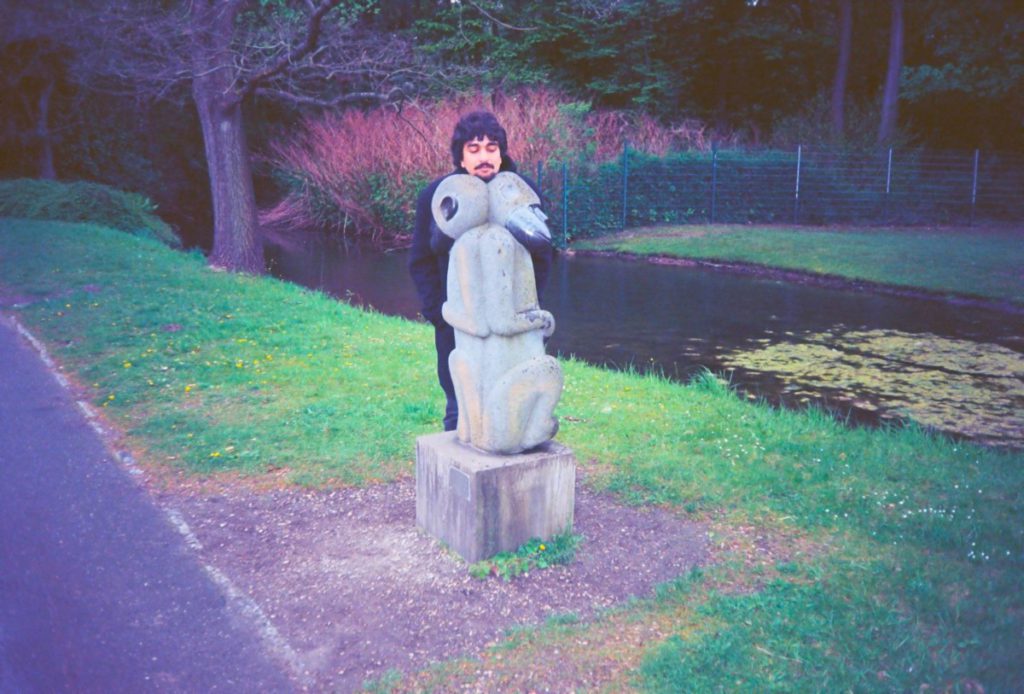 An Indian man in a park standing behind a stone sculpture placing his head on top
