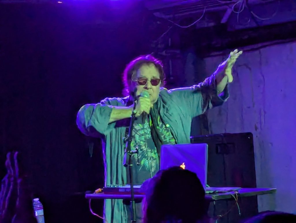 Edward Ka-Spel gestures with his hand while singing in concert with the Legendary Pink Dots