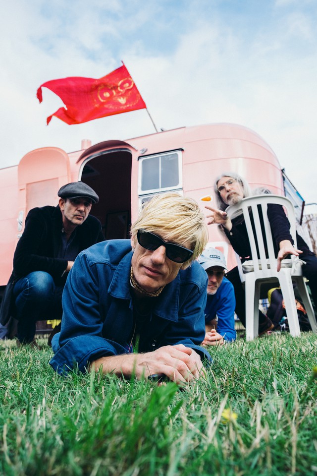 Photo of the band Kula Shaker with Crispian Mills laying on the grass in a blue shirt with sunglasses, the other members behind him and a pink building with a red flag behind them.