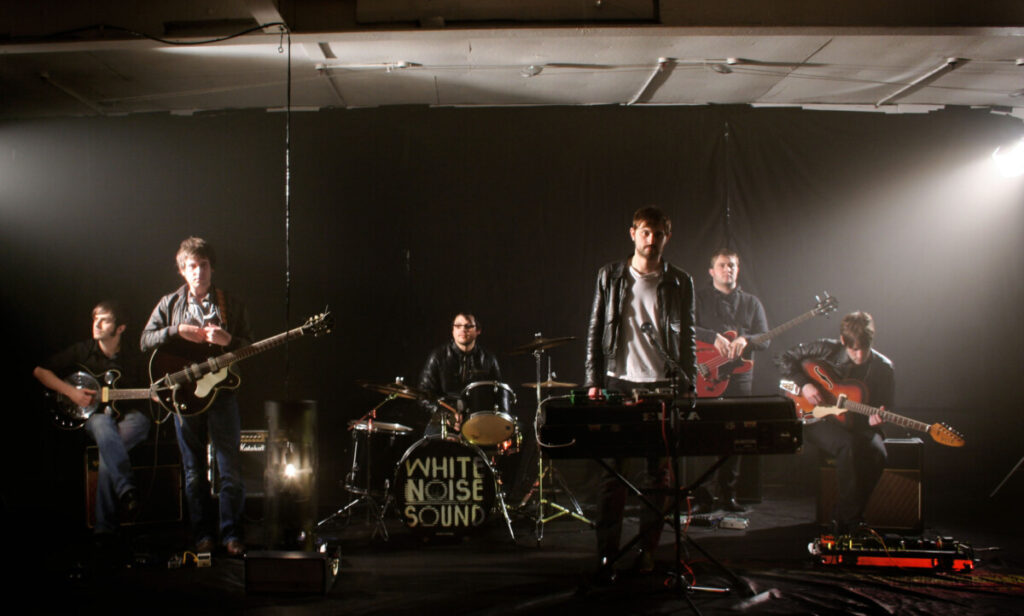 The band, White Noise Sound, in a dark rehearsal room looking at the camera.