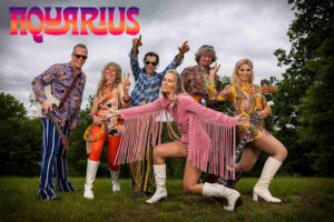 Photograph of six members of band Aquarius dressed in 60s garb and poing in a green field with trees