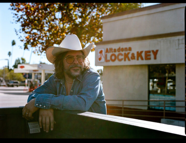 Man with long hair and beard wearing cowboy hat and leaning on a rail.