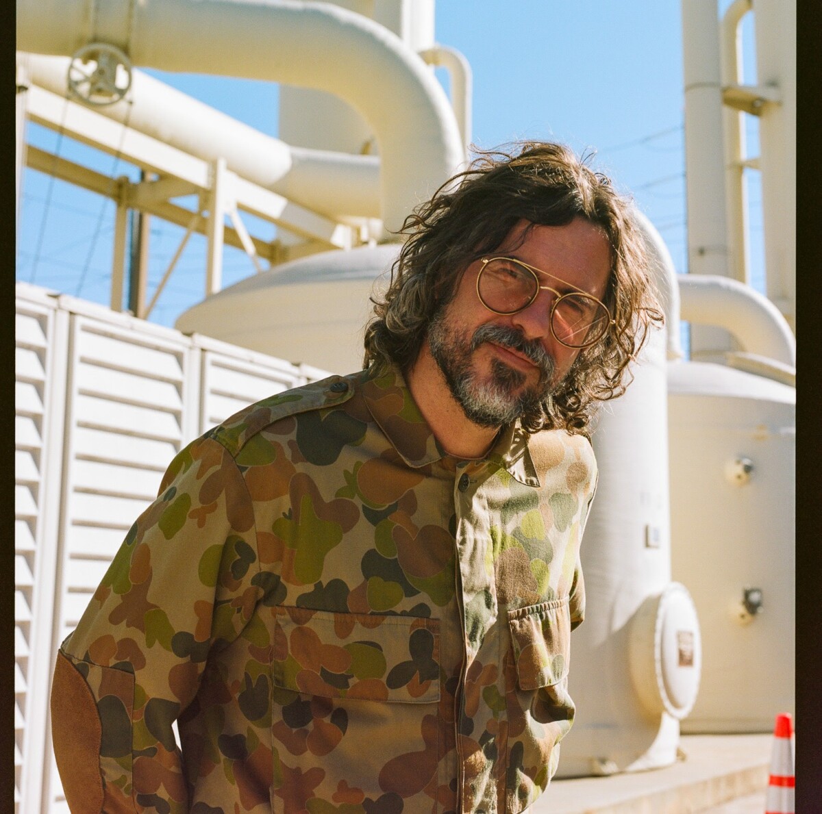 Man with long hair and a beard wearing long-sleeved army fatigues standing in front of power plant