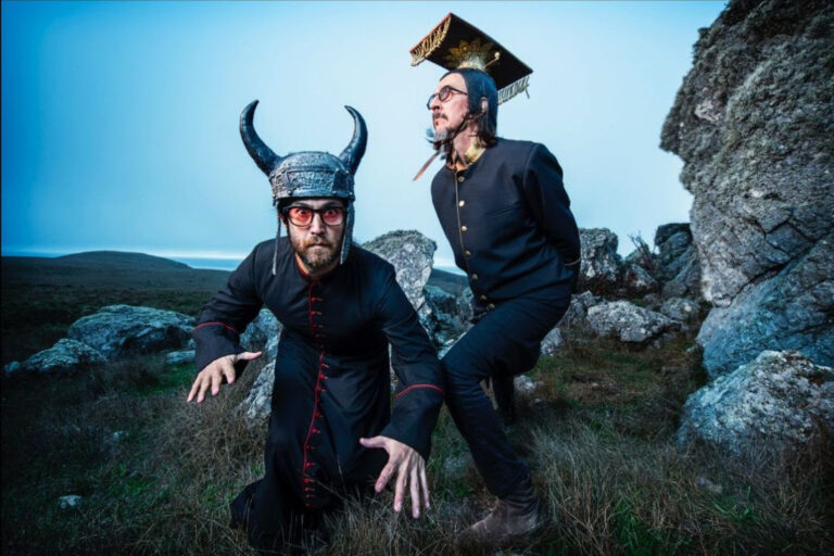 Two men standing near large rocks. One of them has iron horns on and is facing the camera in attack mode while the other looks off into the distance.