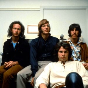 The four members of The Doors sitting on a bed with Jim Morrison in front in what looks like a motel room.