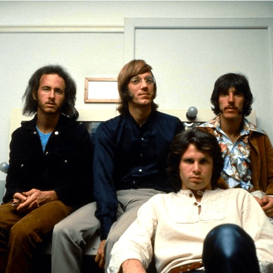 The four members of The Doors sitting on a bed with Jim Morrison in front in what looks like a motel room.