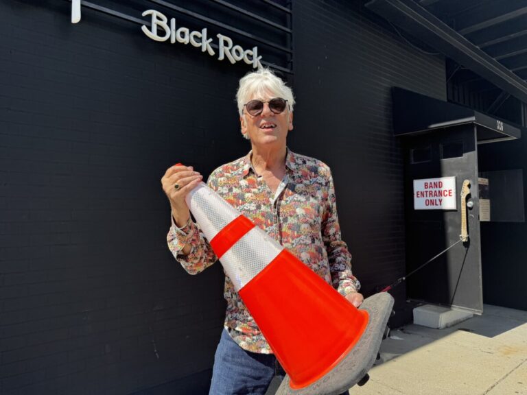 Robyn Hitchcock holding a traffic cone in front of a black building