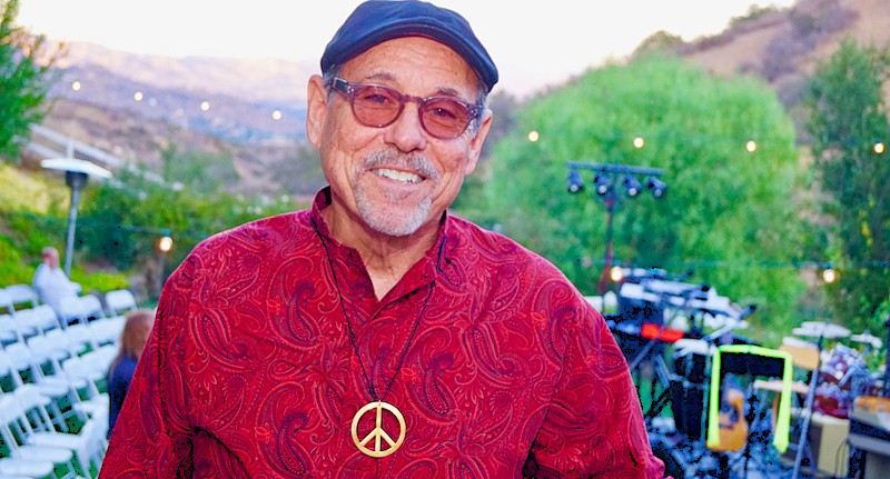 Older man in red shirt, blue cap, pink-tinted glasses and peace sign medallion around his neck outside with green hills in background