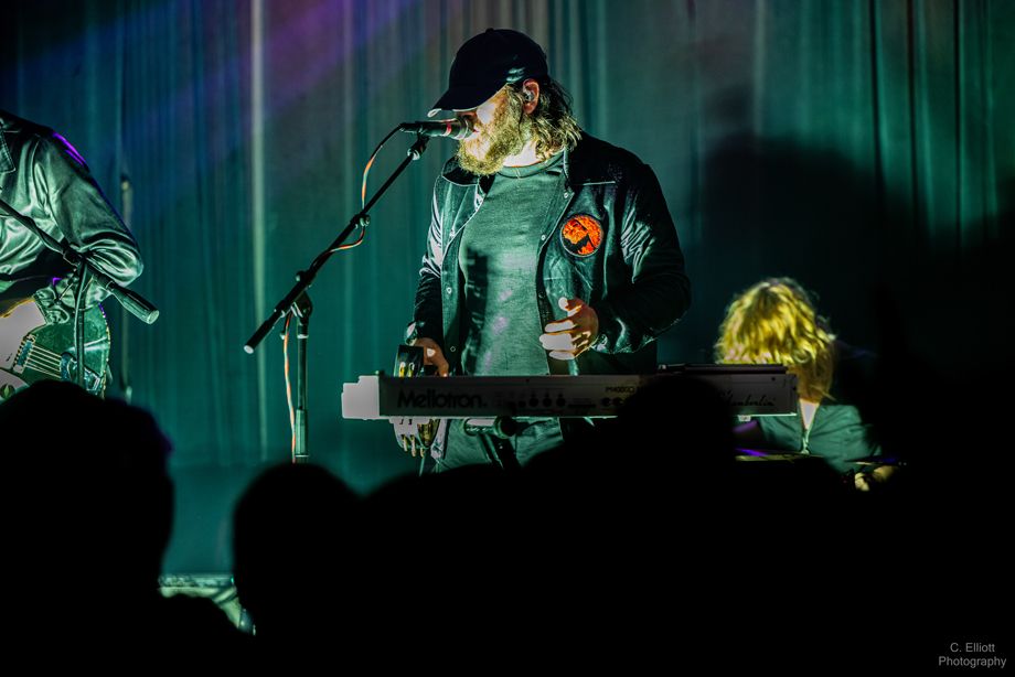 Alex Maas in dark cap and jacket holding tamborine in one hand and a microphone in the other