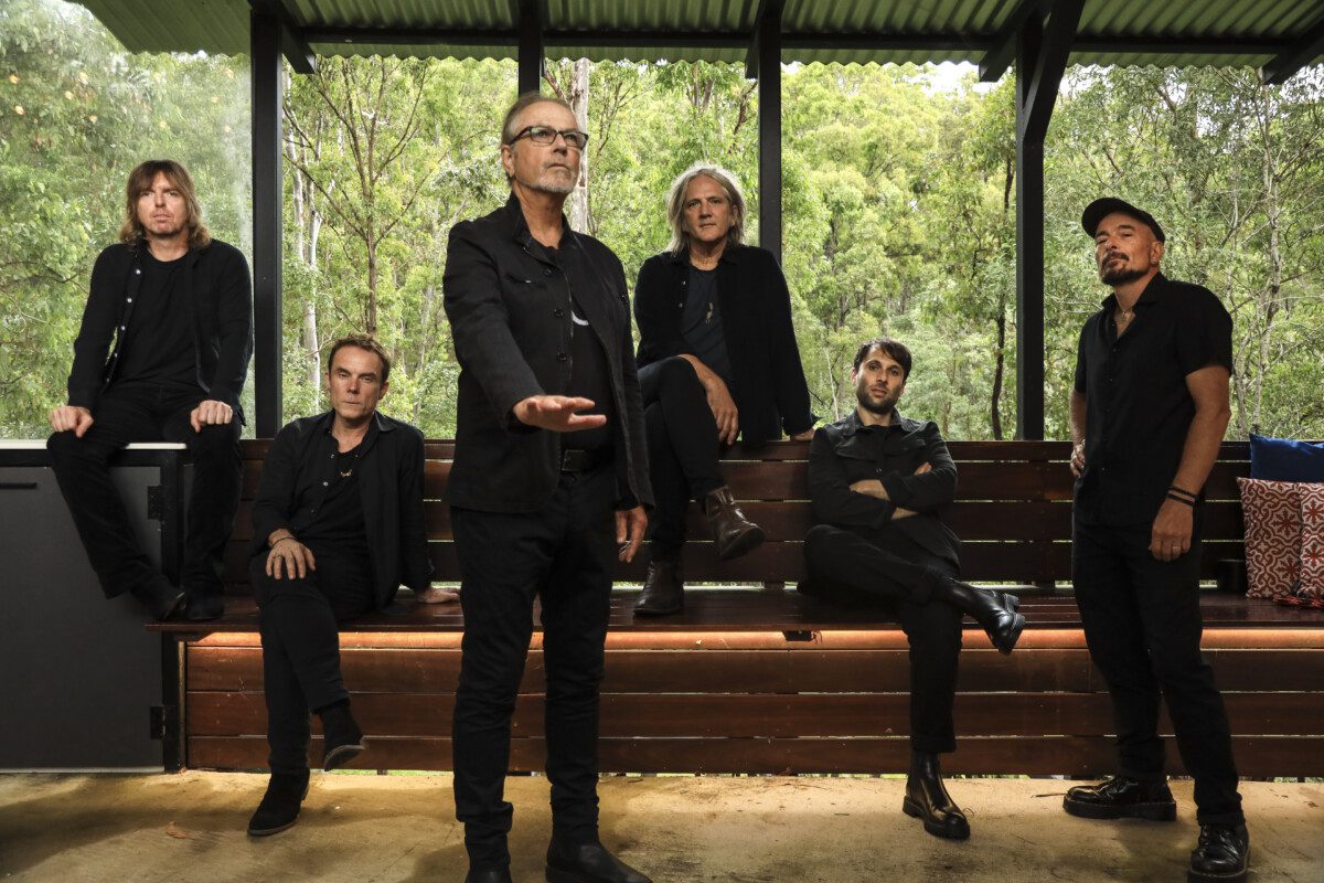 The six members of The Church wearing black while two of the men stand and the four others sit in some type of arbortoreum